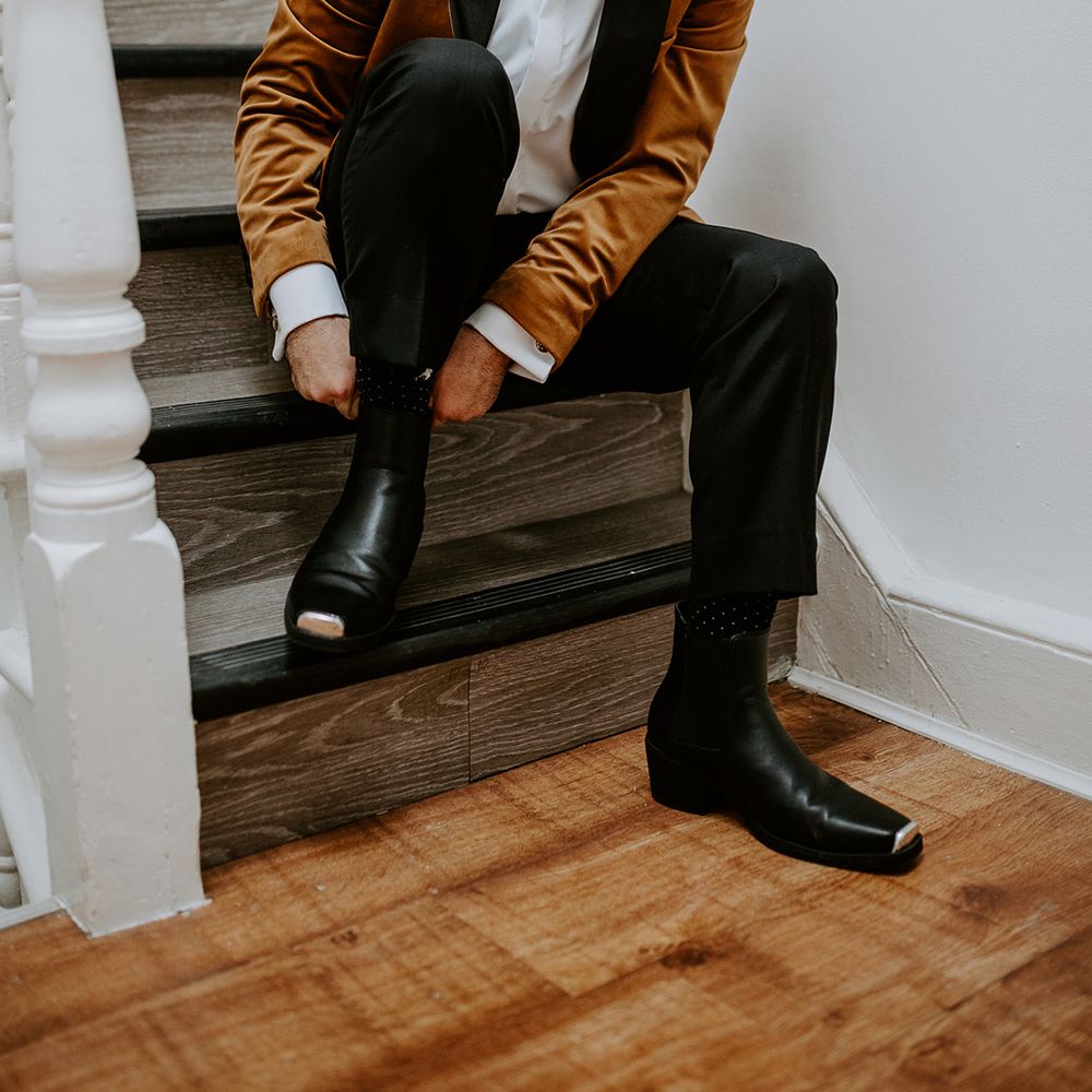 Groom in burnt orange velvet blazer puts on black boots while sitting on staircase