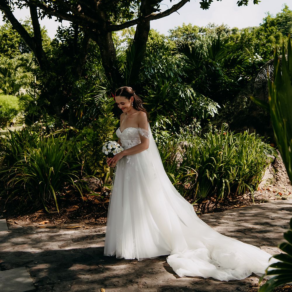 Bride in off the shoulder lace wedding dress with sweetheart neckline and lace floral details and cathedral length veil holding white tropical bouquet 
