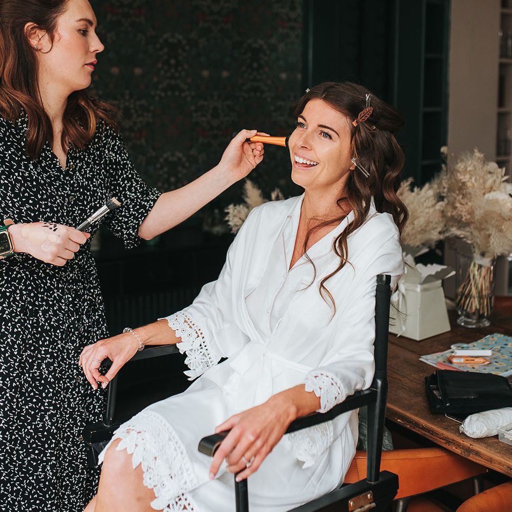 Bride in white robe with crochet trim sits getting her makeup done for her wedding day 