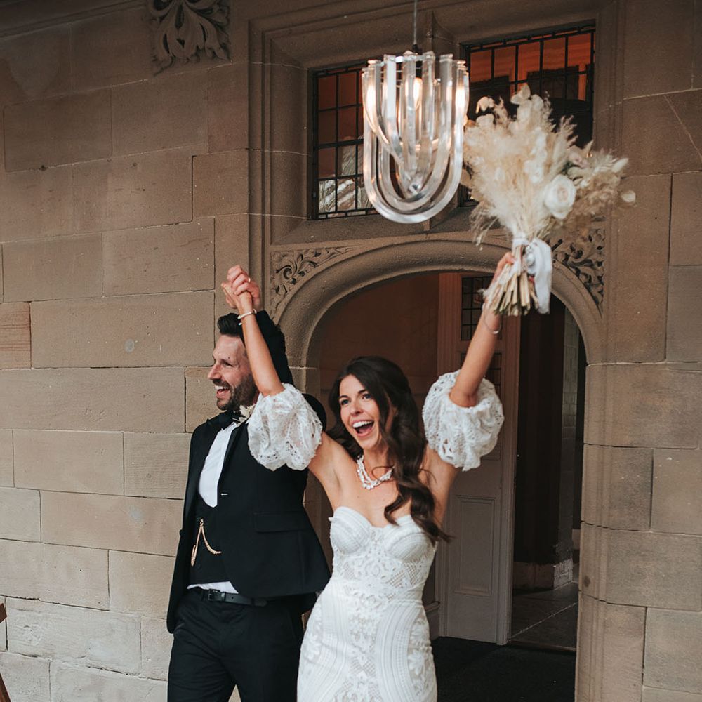Groom in black tuxedo enters the wedding reception at Hampton Manor with the bride in detachable sleeve lace gown 