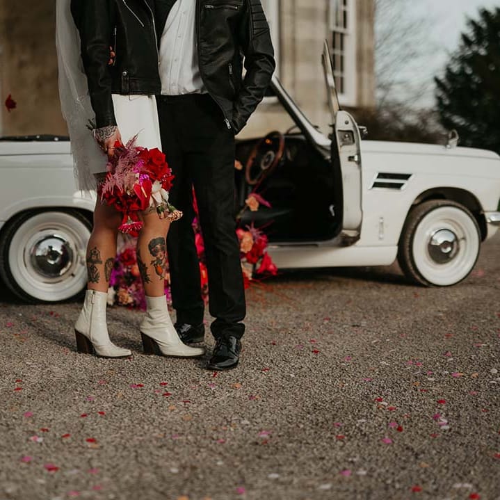 Bride in blazer style wrap around short wedding dress with delicate fringe detailing and personalised, embroidered black leather jacket  with white patent heeled ankle boots with black soles and groom in white shirt, black bowtie, black suit trousers and slim fitting black leather jacket standing by off white classic convertible wedding car