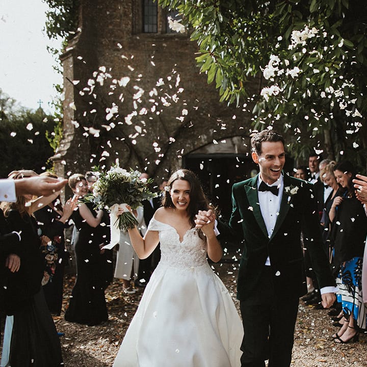 Bride in off the shoulder lace wedding dress walking with the groom in a green velvet tuxedo walk out of their ceremony to confetti 