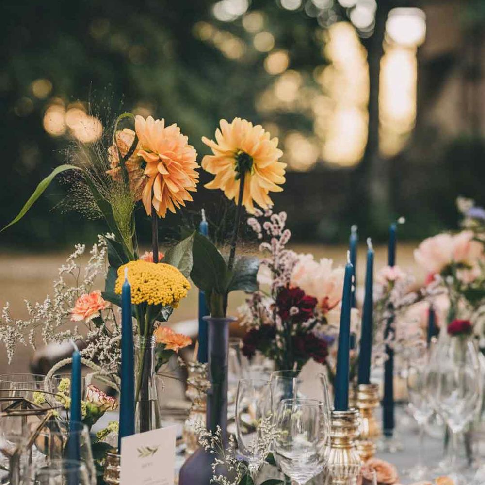 Blue tapered candles and yellow flower stems in vases and bottles on rustic, natural wedding tablescape 
