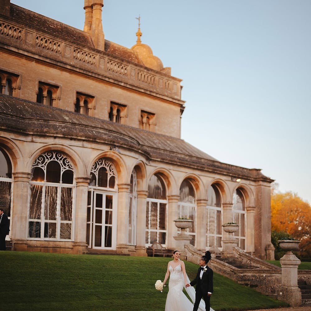Bride in lace sleeveless v neck Enzoani wedding dress with tulle finish and church length veil holding white rose bouquet walking hand in hand with bride in black snakeskin print tuxedo with black bowtie and white rose boutonniere on the grounds of Grittleton House wedding venue  