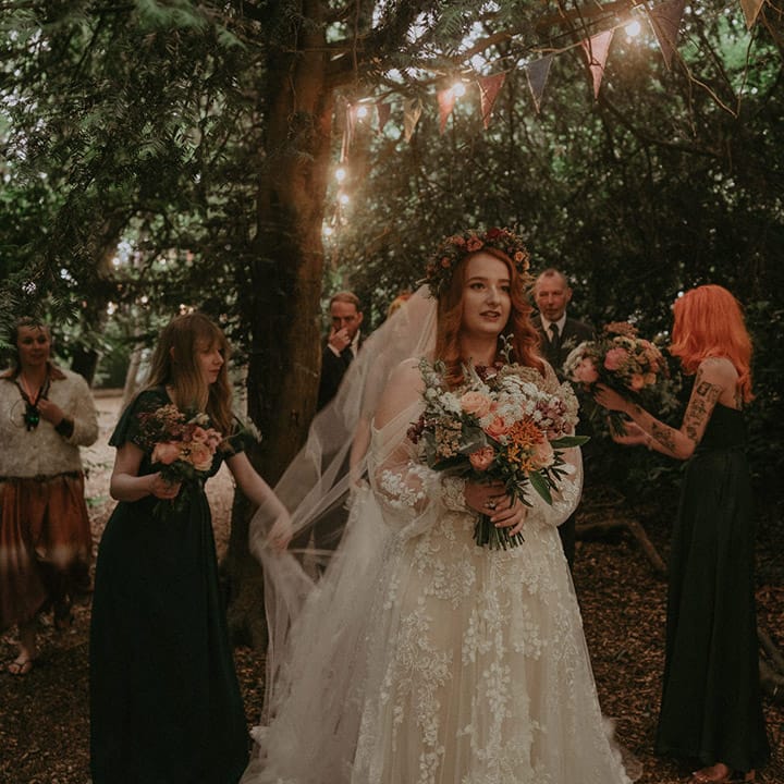 Bride in floral lace boho wedding dress gets ready to walk down the aisle with the bridesmaids in green bridesmaid dresses and the father of the bride 