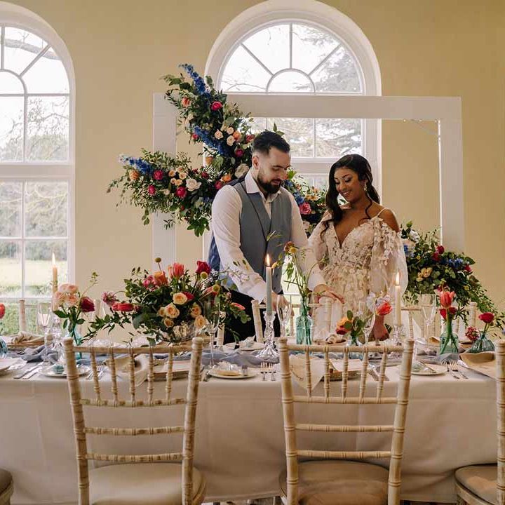 Bride in v-neck long sleeve shoulder cut out 3d appliqué wedding dress with flecks of gold standing with groom in cornflower blue waistcoat and black suit trousers by classic wedding tablescape with white wedding tablecloth, cornflower blue wedding table runner, bright blue glass bottles with dried flowers, pastel tapered candles and spring floral arrangements with suspended spring floral decor 