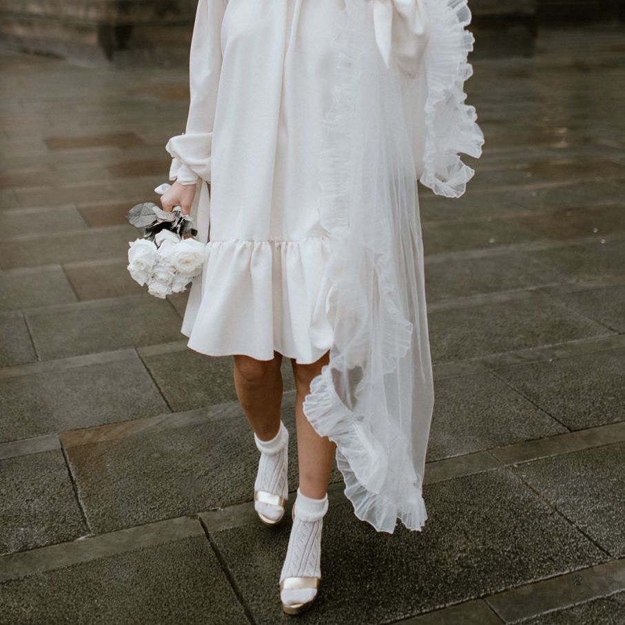 Stylish bride with short fringe, red lipstick and gold Gucci shoes wearing a registry office wedding dress holding a white rose bouquet 