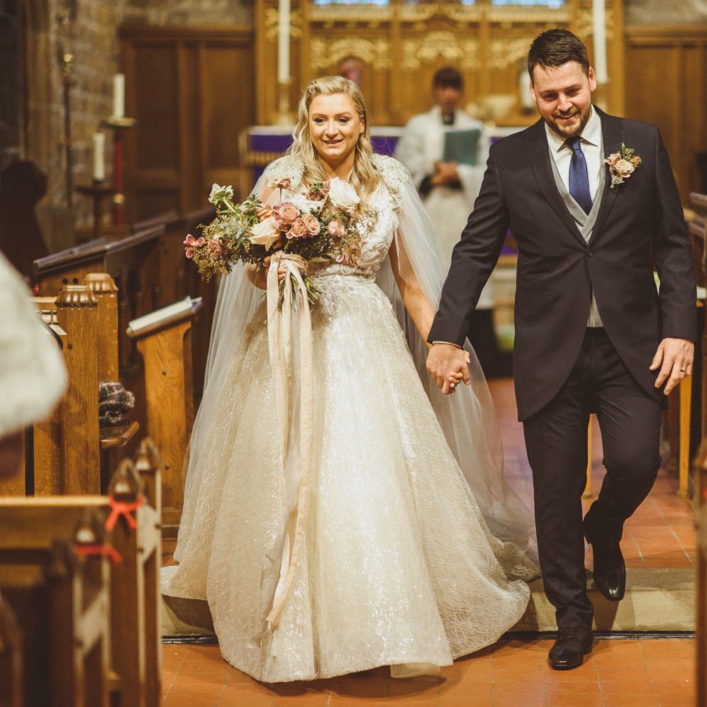 Groom in a navy morning suit holding his brides hand in a princess wedding dress walking up the aisle as husband and wife 