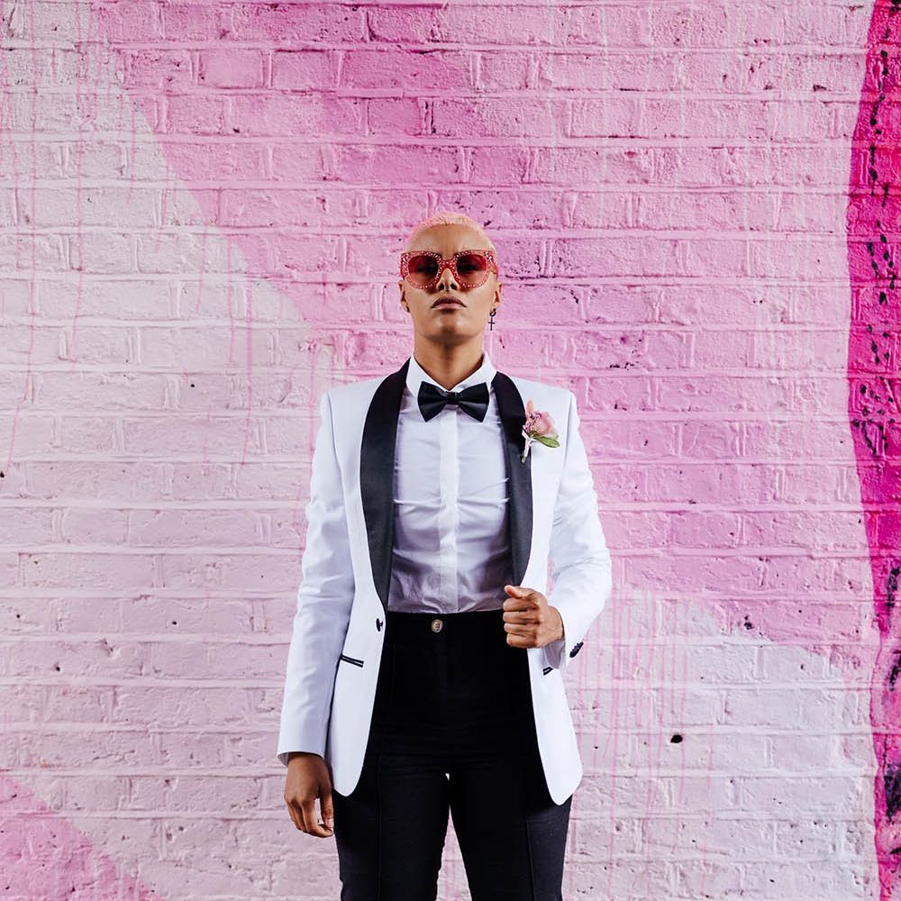 Bride in a white tuxedo jacket with black bow tie standing in front of a pink wall 