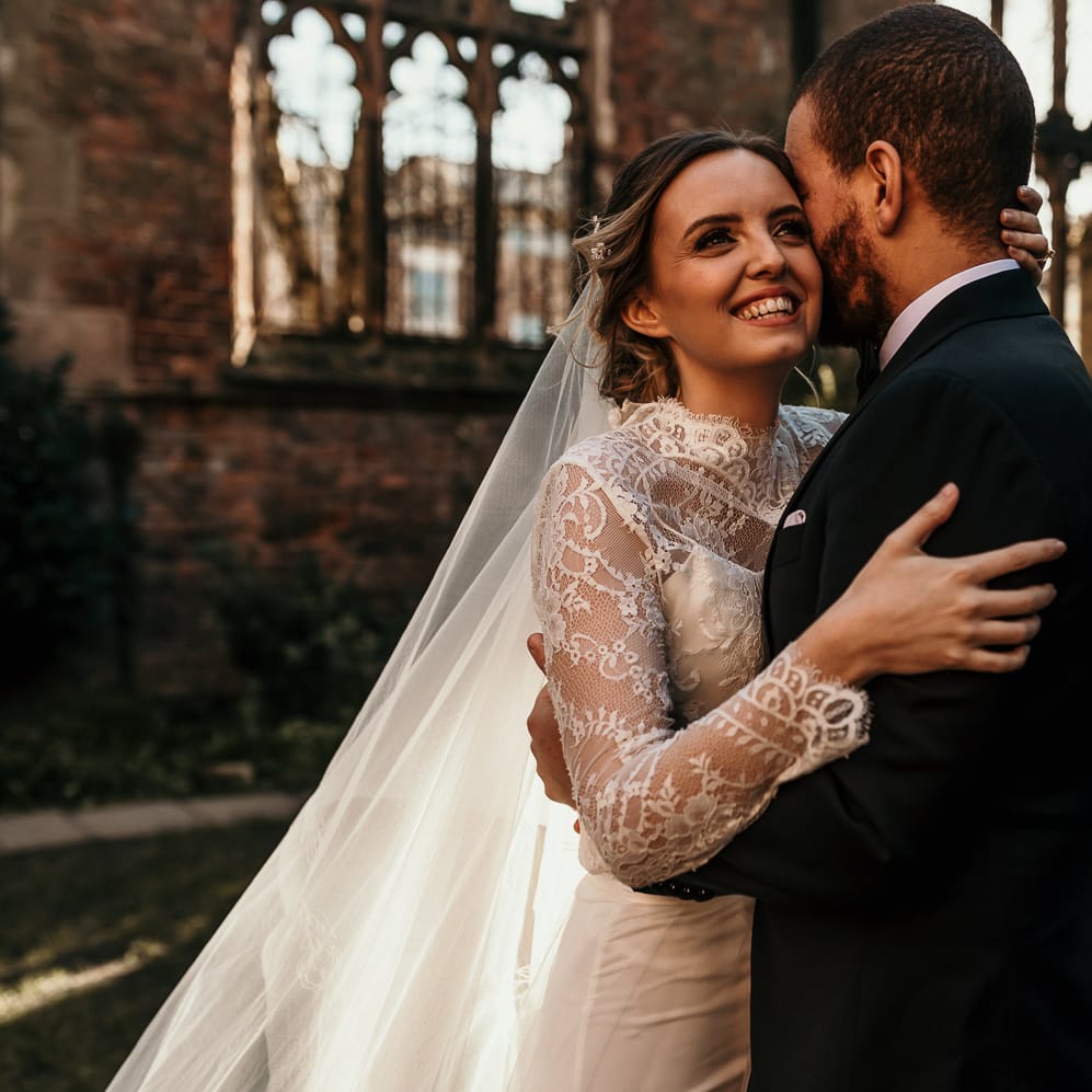 Beautiful bride in a lace wedding dress and cathedral length veil smiling as she embraces her husband in a black suit 