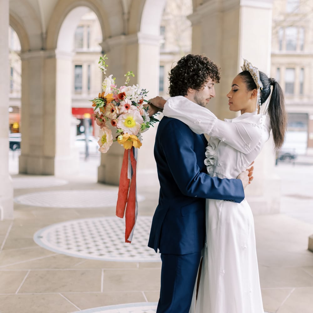 Bride & groom embrace outdoors as bride holds floral bouquet tied with ribbon