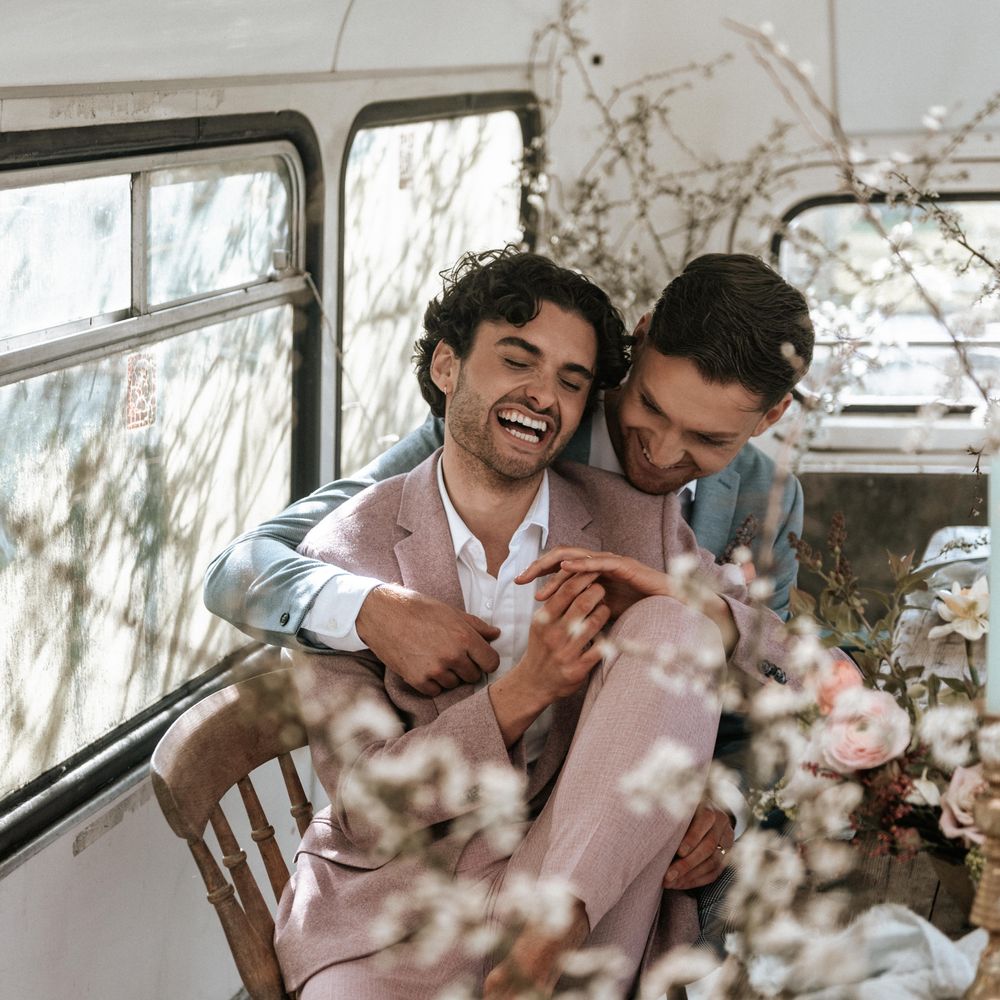 Grooms sit beside rustic and pastel coloured floral bouquets with wooden twisted vines 