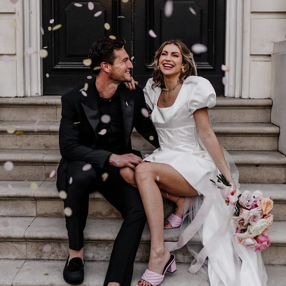 Modern luxury wedding day moment with bride and groom sitting on some steps as confetti is thrown over them
