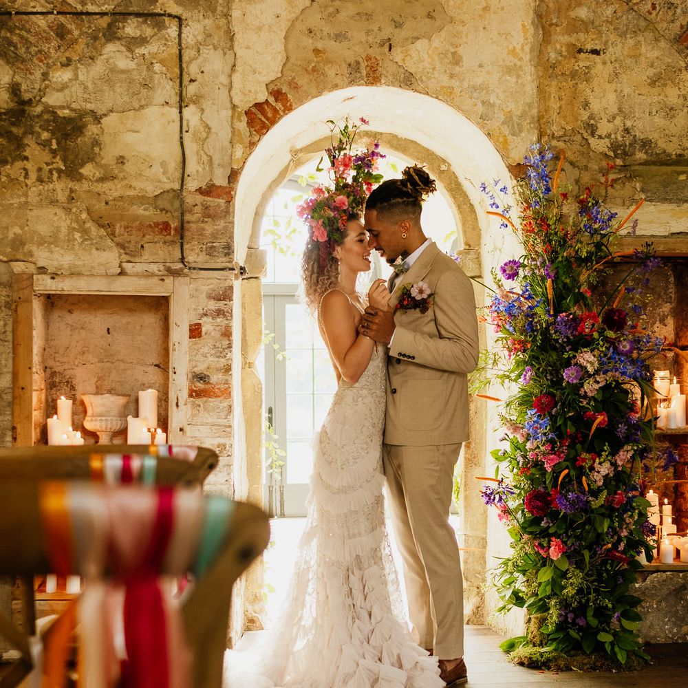 Bride in a layered ruffle skirt wedding dress and colourful flower crown standing at the altar with her husband in a beige suit next to a vertical flower arrangement 