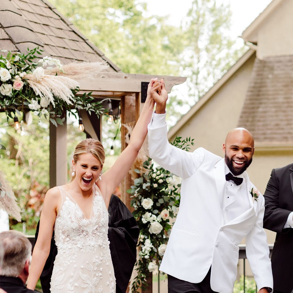 Bride in white embroidered Made With Love wedding dress and groom in white tuxedo jacket hold hands in the air in joy after being married at backyard wedding