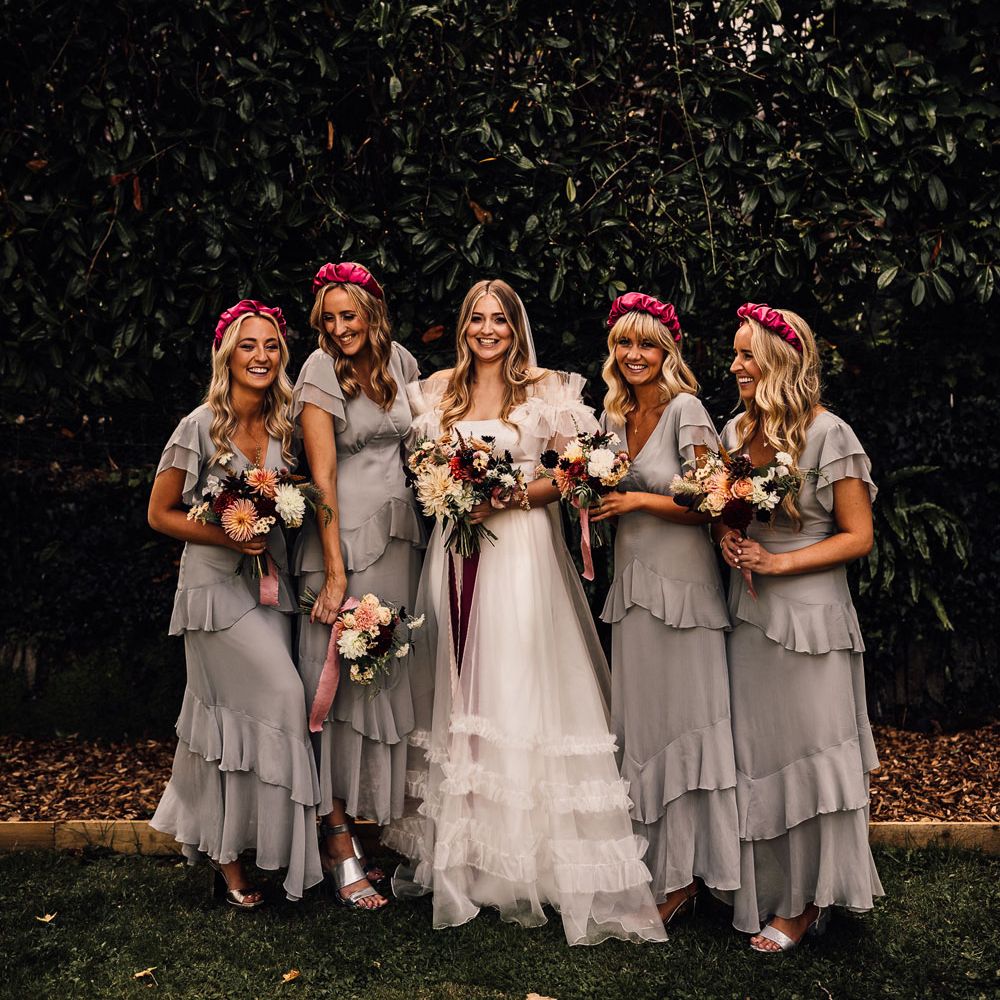 Bridesmaid in white Halfpenny London Mayfair dress holding mixed bridal bouquet stands in the middle of bridesmaids in grey ruffle bridesmaid dresses and pink headbands before church ceremony in Cornwall 