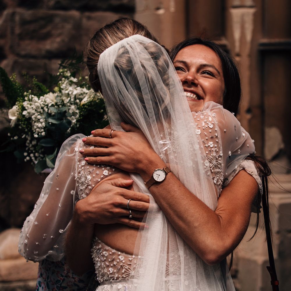 Bride in open back Julie Vino embroidered wedding dress and veil hugs smiling wedding guest after church wedding ceremony