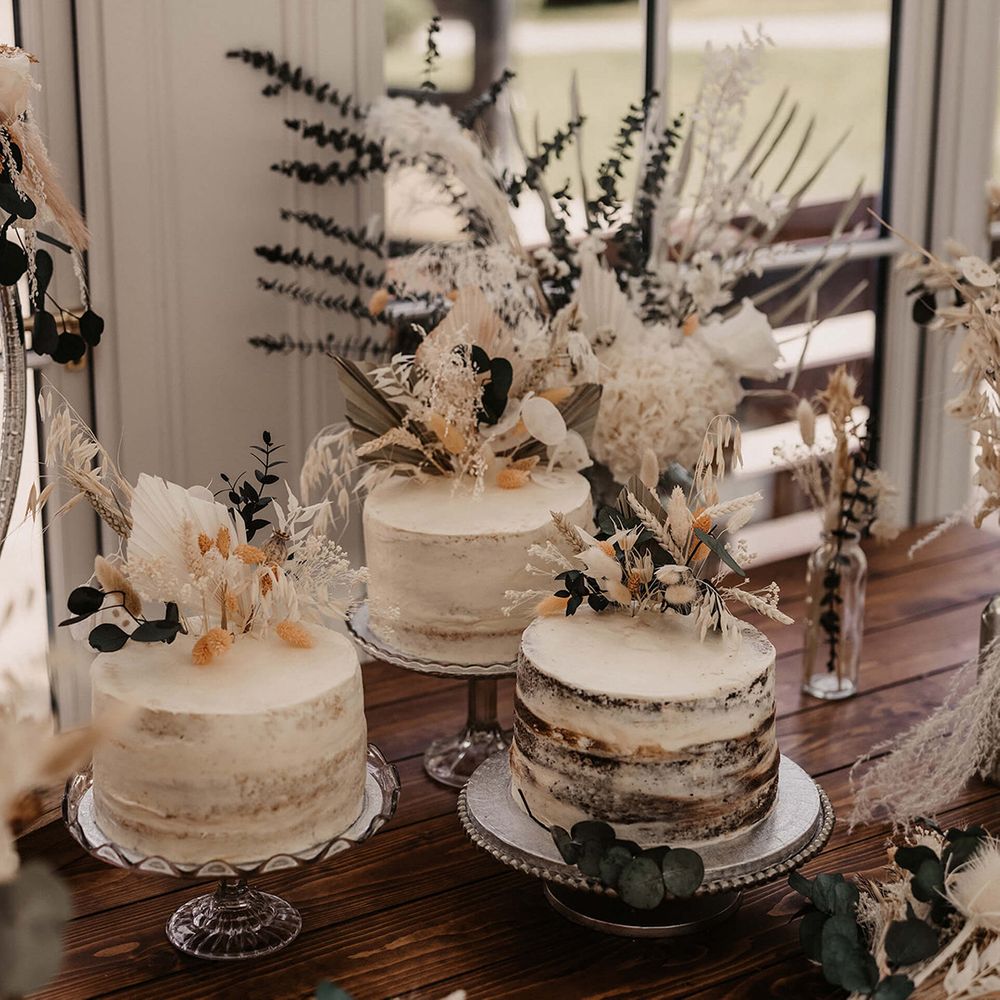 Three naked wedding cakes complete with white frosting next to dried florals