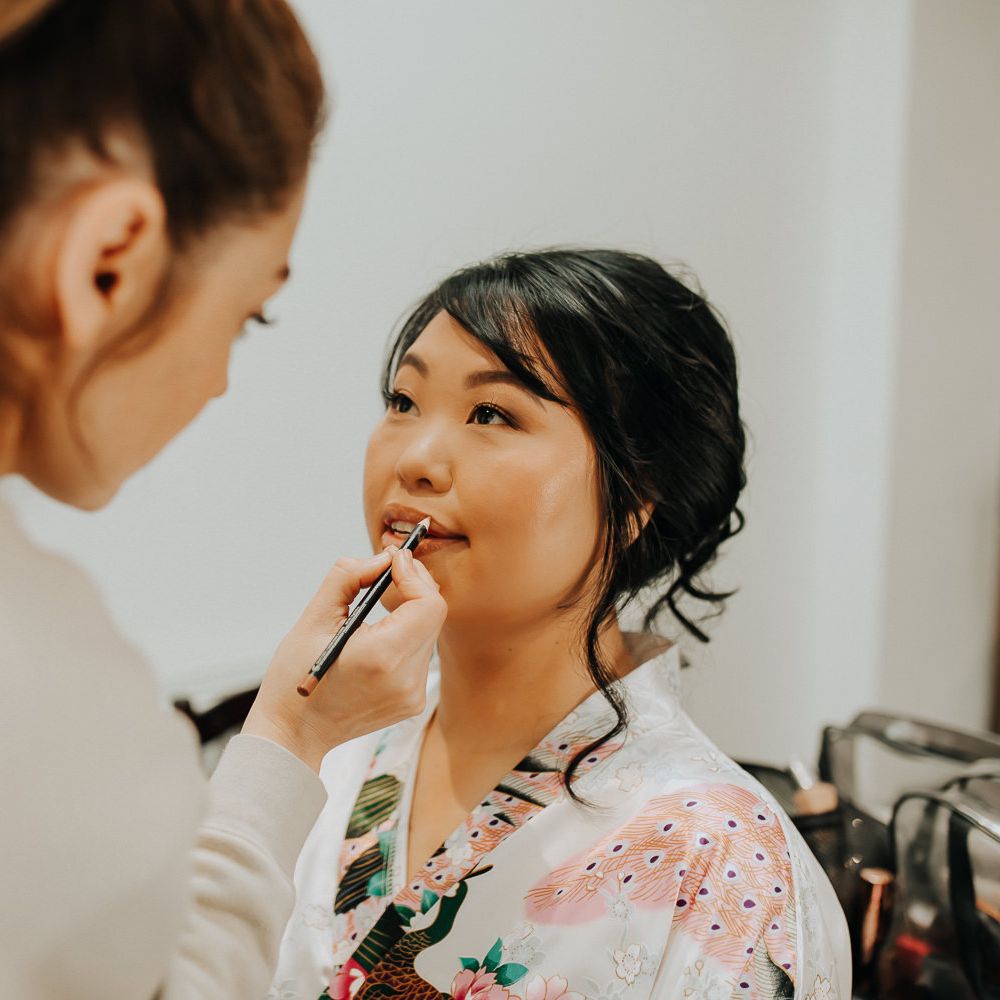 Bride in patterned silk kimono dressing gown sits having her lips lined before wedding 