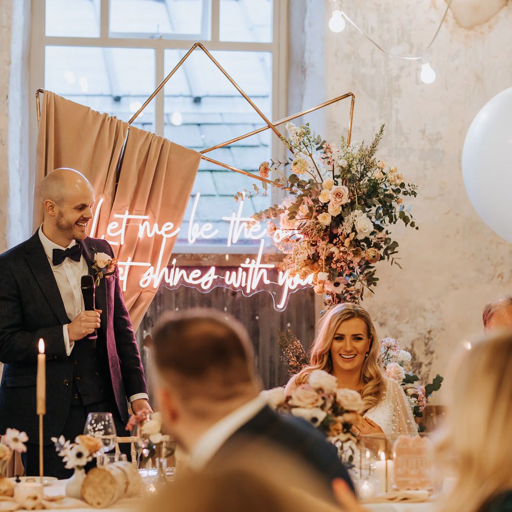 Groom in navy suit holds microphone whilst making speech during wedding reception with copper frame wedding arch and neon sign