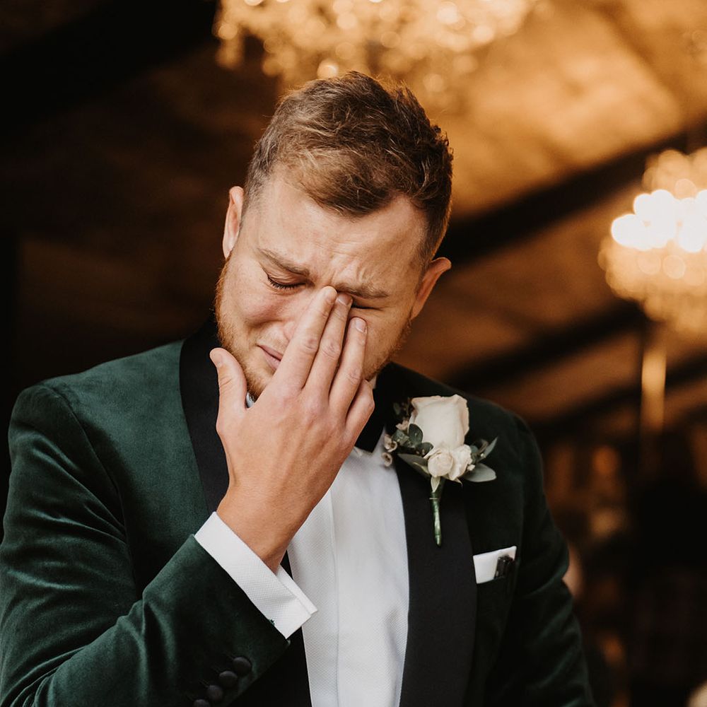 Groom in velvet green suit jacket and white rose boutonnière cries as he sees his bride 