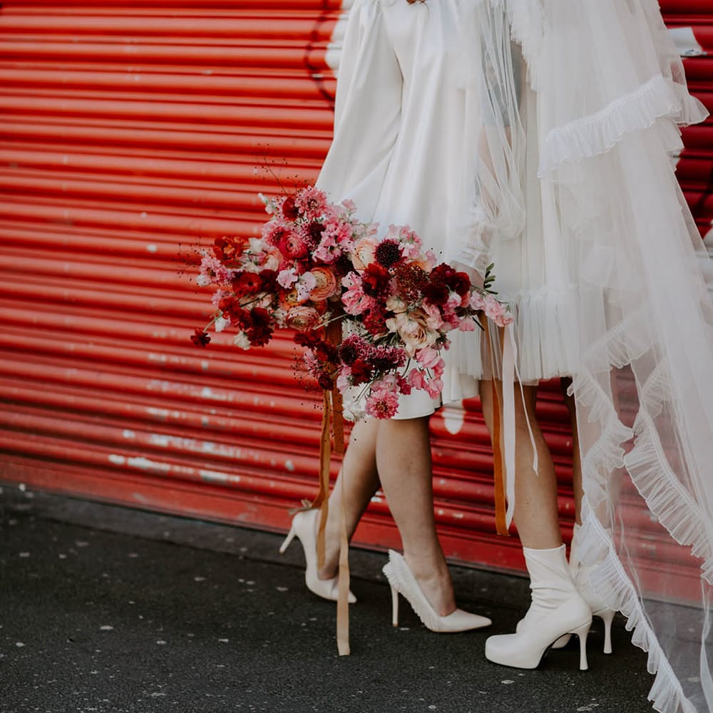 bride in short wedding dress long veil and boots wipering into her brides ear in a short dress and berret hat as she holds a red and pink bouquet 