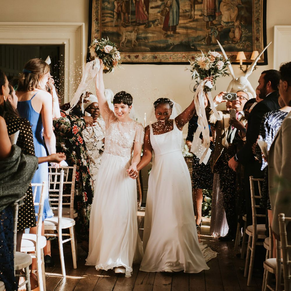 LGBTQI+ wedding ceremony at Elmore court with two brides descending up the aisle as wife and wife waving their bouquets in the air 