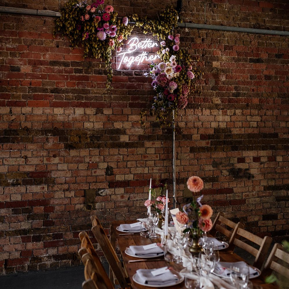 Better together neon wedding sign surrounded by green crops and pink wedding flowers on an exposed brick wall 