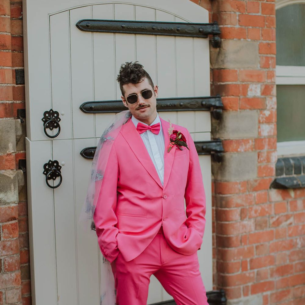Stylish groom in a pink two piece suit with bow tie, sunglasses and embroidered wedding veil 