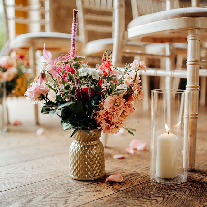 White pillar candles in glass cases beside bright floral bouquets in gold vases alongside petals line the aisle at The Barn At Upcote