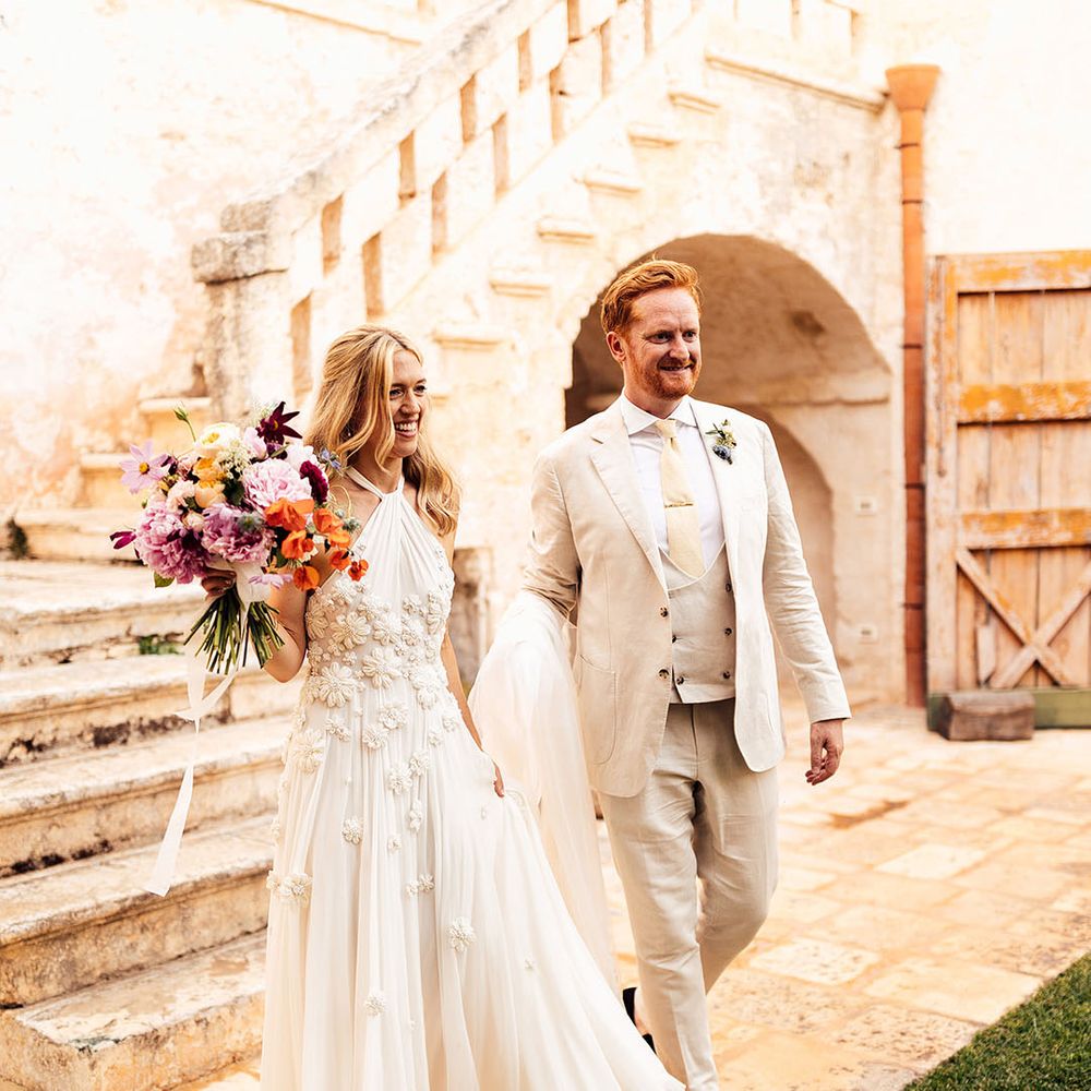 Groom in a three piece cream wedding suit walking with the bride in a halter neck flower wedding dress 