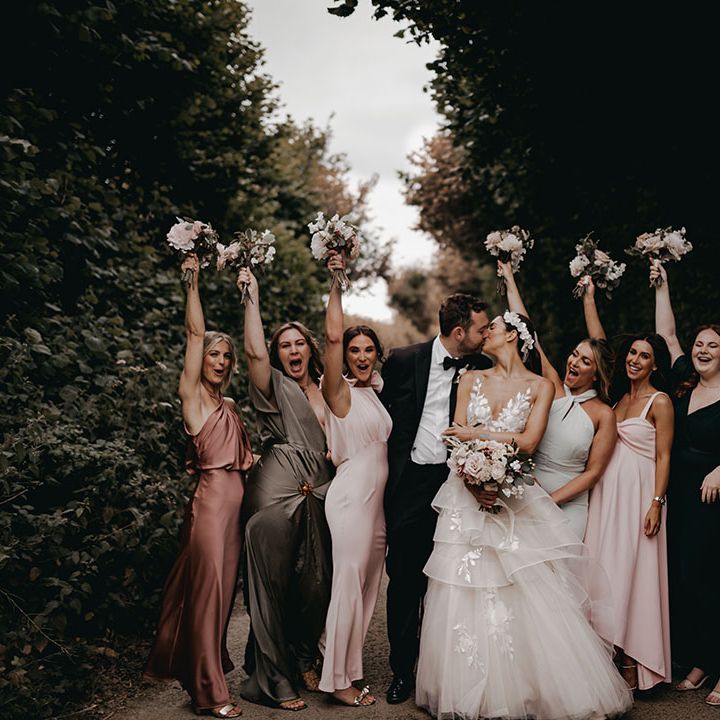 The bride and groom kiss as they pose with the bridesmaids in different coloured and mismatched dresses