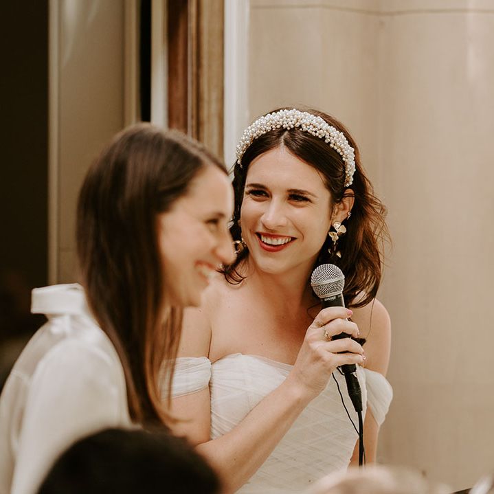 The bride wearing a pearl headband and statement earrings stands with a microphone to deliver a speech 