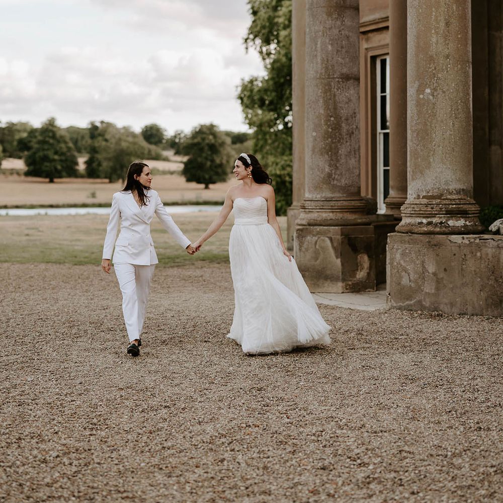 The bride in an off the shoulder wedding dress and bride in suit walk holding hands at the Wilderness Reserve wedding venue 