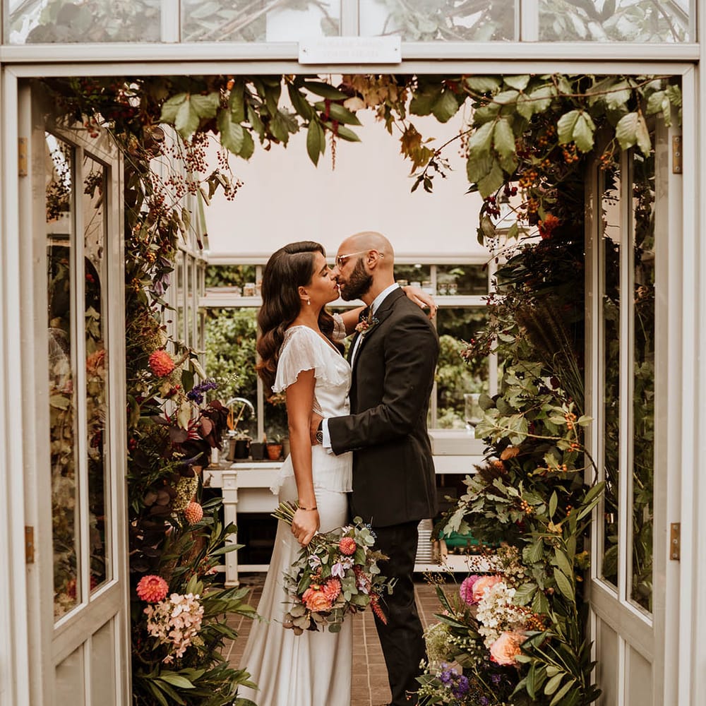 Bride in v neck satin wedding dress with sheer butterfly sleeves and ruffled layer on top kissing groom in classic black tuxedo with hand painted silk cravat and handkerchief at Le Manoir Aux Quat Saisons glasshouse venue