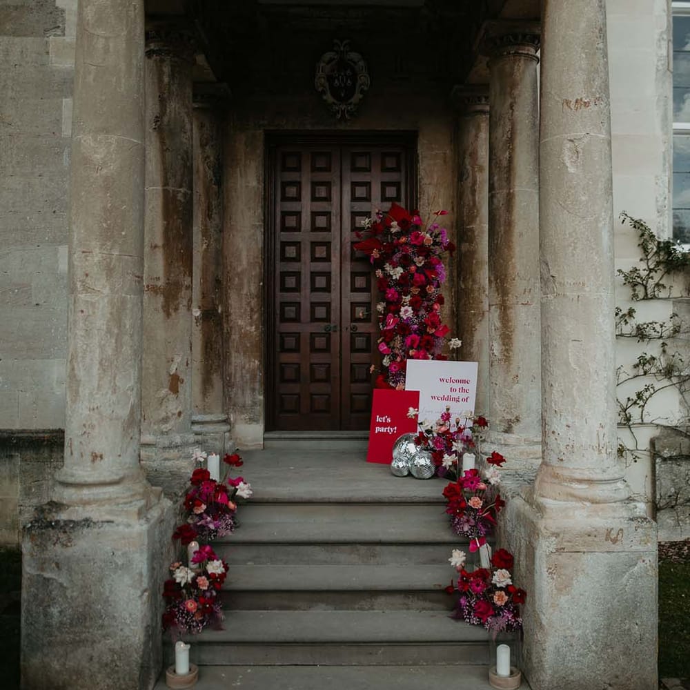 Large wedding floral decorations with hot pink, bright pink dahlias, orchids, roses, amaryllis, anthuriums and dried flowers on the steps of Elmore Court with disco ball decorations and viva magenta and baby pink retro wedding stationery 