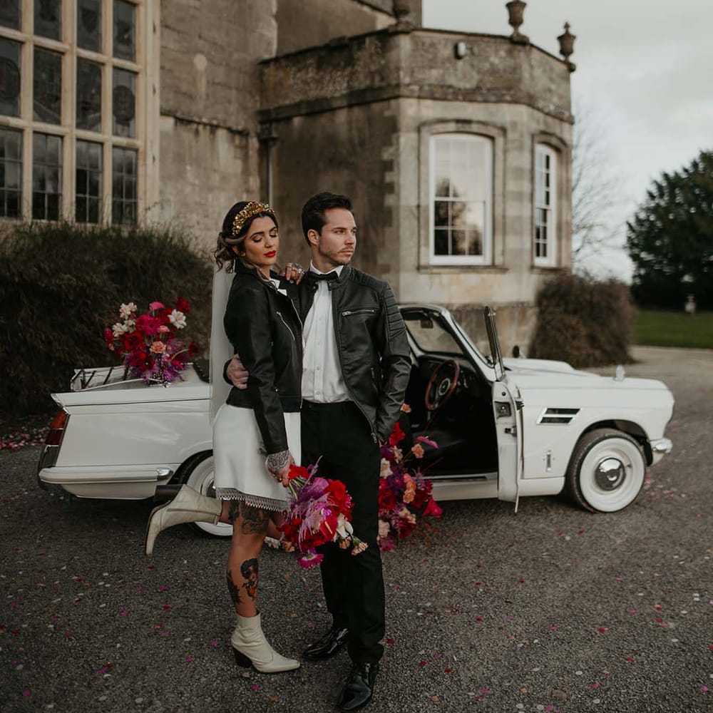 Groom in white shirt, black bowtie, black suit trousers and slim fitting black leather jacket embracing bride wearing blazer style wrap around short wedding dress with delicate fringe detailing and personalised, embroidered black leather jacket by off white classic convertible wedding car