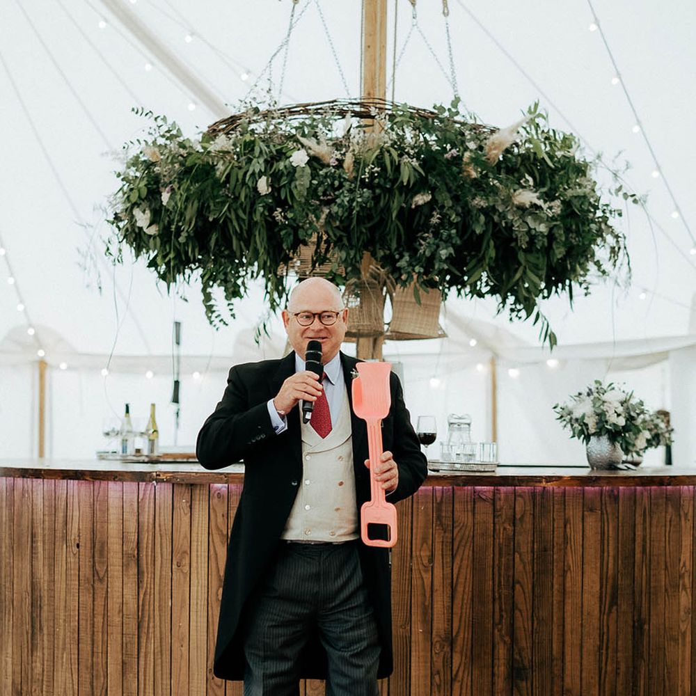 Wedding guest gives speech in marquee beside green foliage floral installation and wooden bar