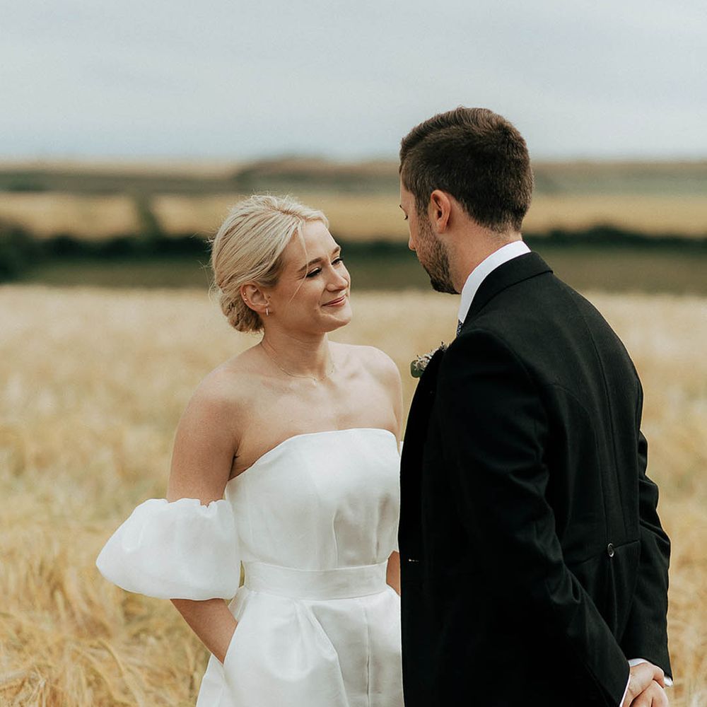 Bride wears Jesus Peiro wedding dress and places her hand in the pockets as she stands in golden fields with her groom