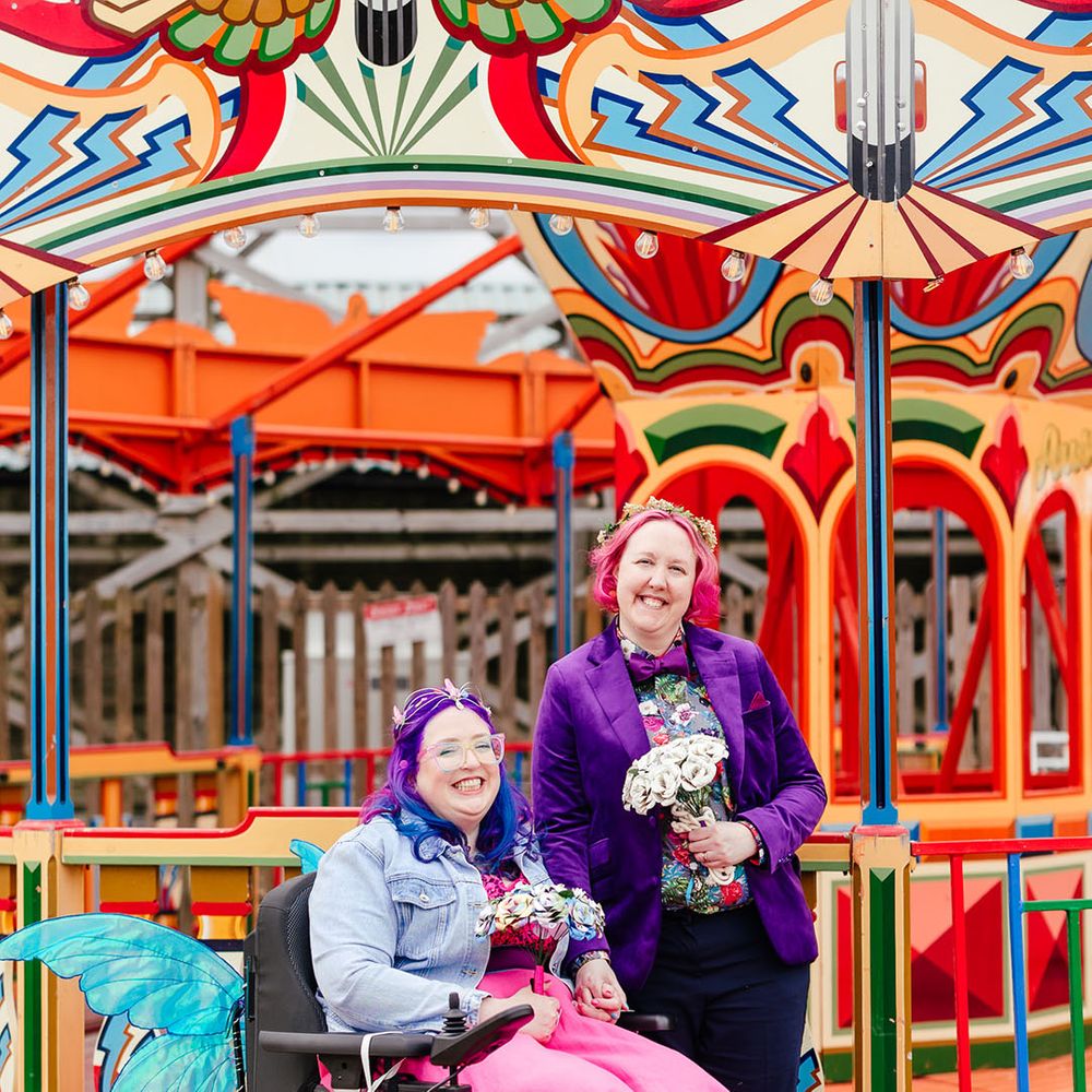 Bride in wheelchair with sparkly blue fairy wings hung on wearing personalised denim jacket, pink wedding dress and blue fishnet tights with bride in purple velvet blazer and black suit trousers with purple bowtie, pink patterned pocket square and colourful patterned shirt