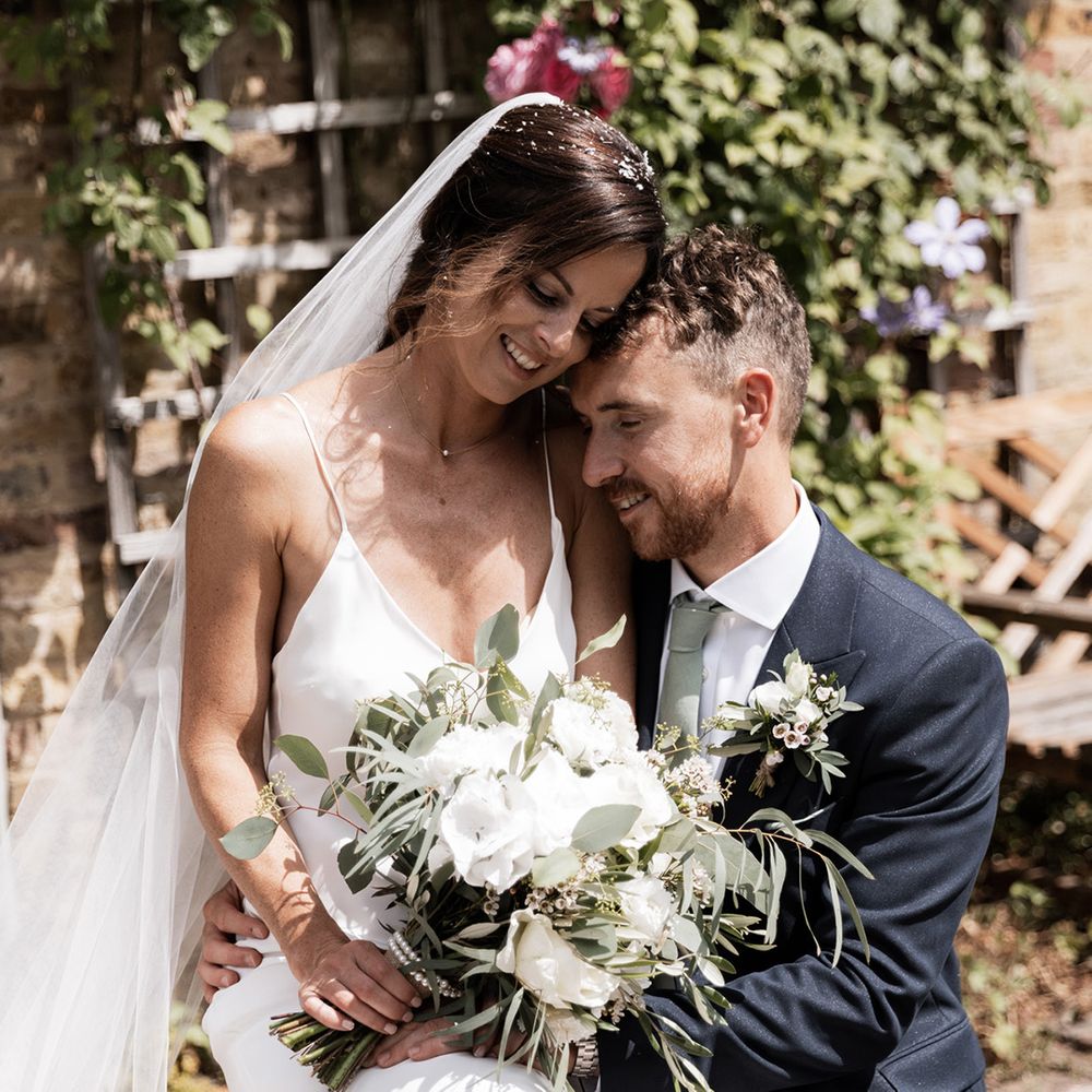 Bride holds white floral bouquet and Grace Loves Lace wedding dress whilst sat beside her groom in double breasted blazer 