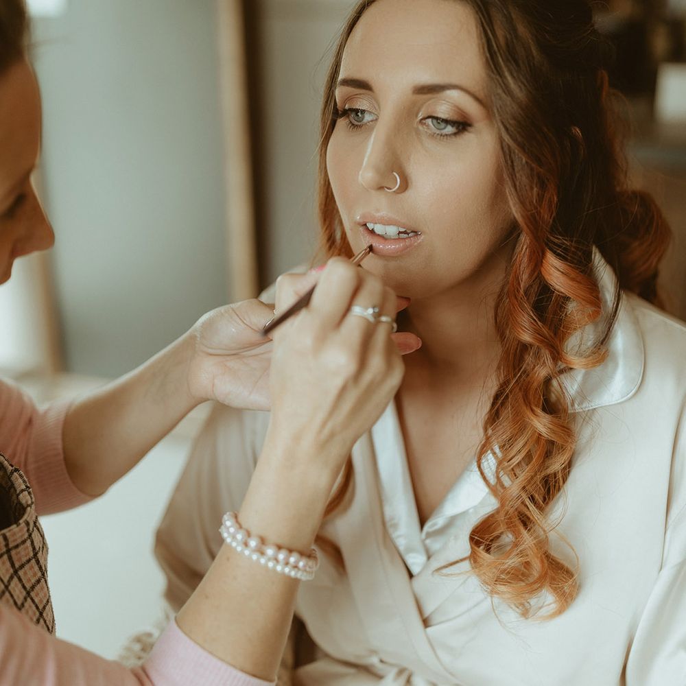 Bride with curled red hair sits in a cream satin robe getting her makeup done for her wedding day 