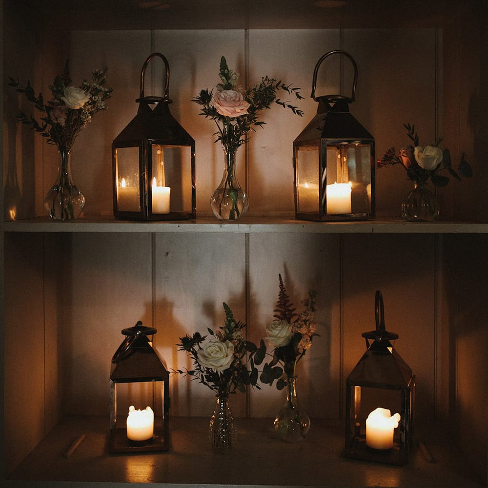 White pillar candles in glass lanterns on a shelf with bud vases full of pink and white roses 