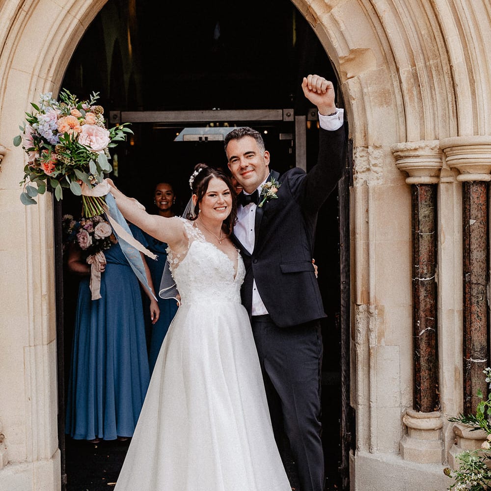 Bride in flower patterned wedding dress puts her arm in the air with the groom to celebrate their wedding with groom in black tie 
