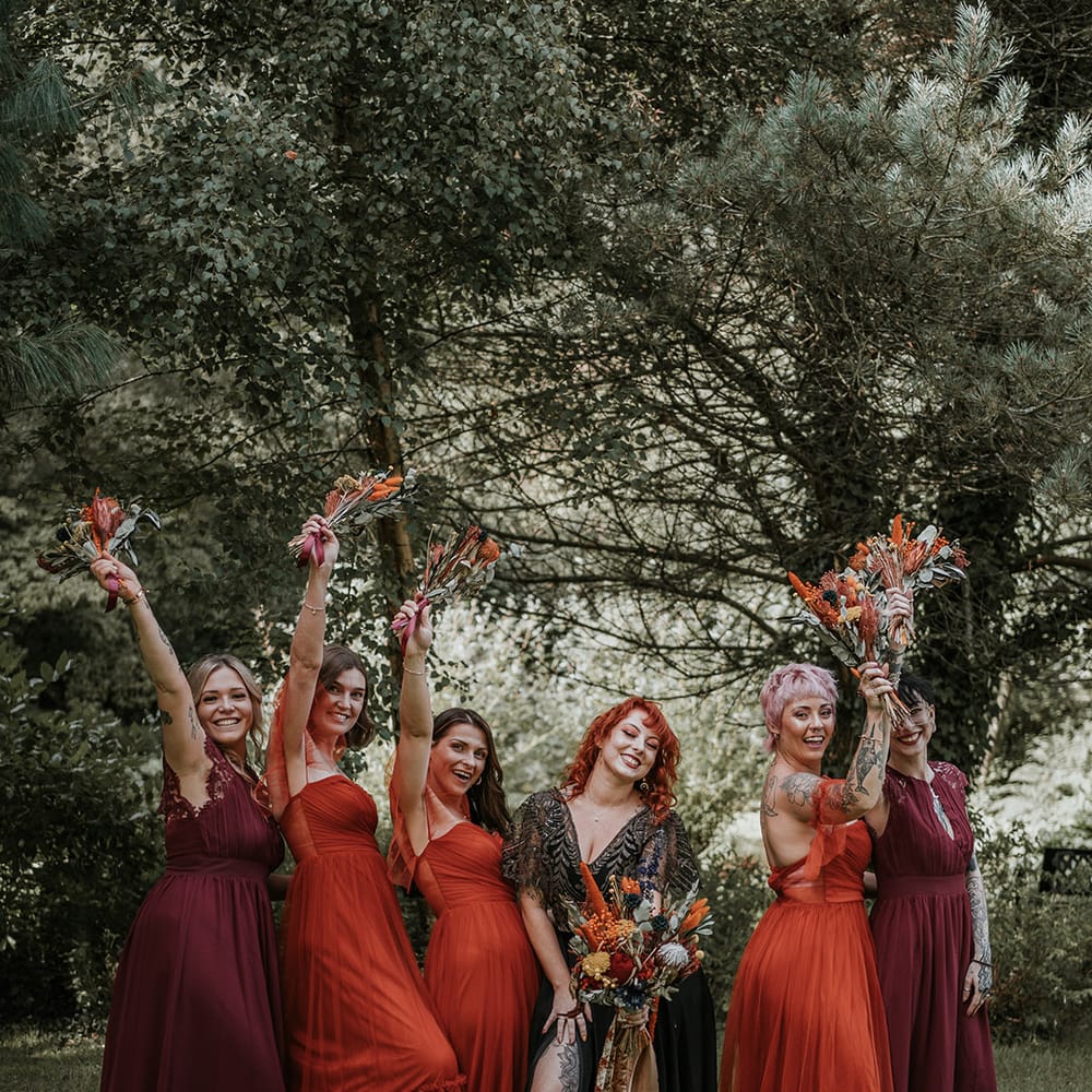 Bride in black dress for wedding in Devon standing with bridesmaids in mismatched burgundy and burnt orange bridesmaid dresses holding burnt orange dried flower bouquets 