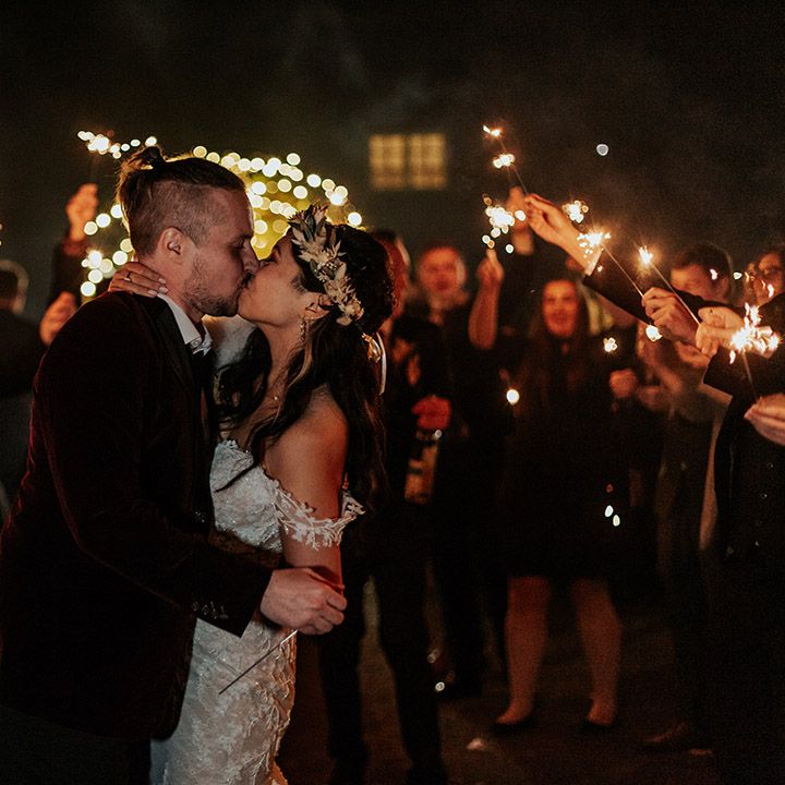 The bride and groom share a kiss at their boho autumnal wedding with the wedding guests waving sparklers for the sparkler send off 