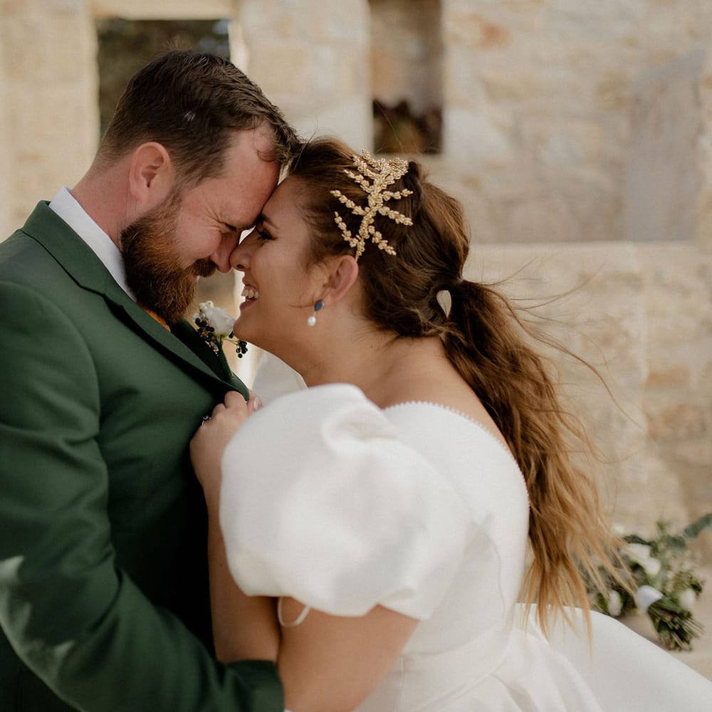 Bride in white satin dress and gold leaf bridal crown touches noses with groom in dark green groom suit at private villa wedding in Greece