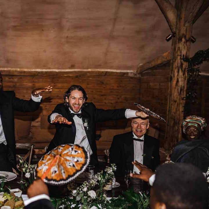 Wedding guests in classic black suits dancing and laughing at Lains Barn holding Ghanaian printed hand fan favours during wedding breakfast 