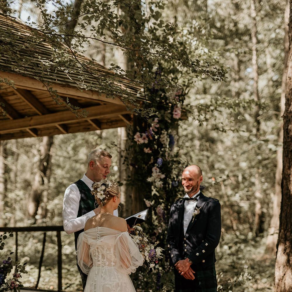 Bride in strapless wedding dress with tulle detachable sleeve standing facing the groom for outdoor woodland wedding ceremony 
