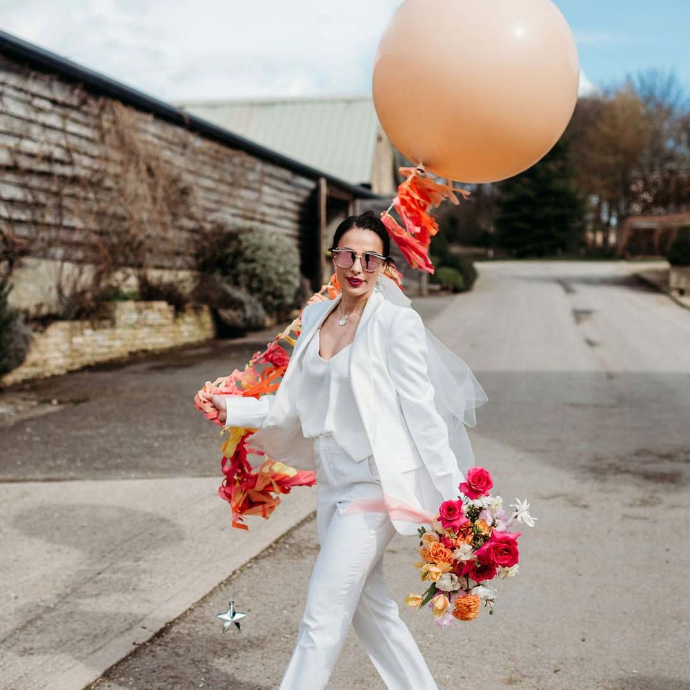 Bride in white bridal suit with closed toe bright green wedding heels holding mixed seasonal floral bouquet and large peach balloon with wedding streamers attached outside of Merriscourt wedding venue